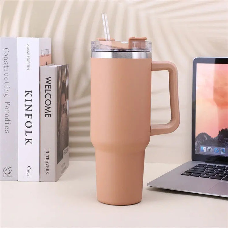 Rostfritt stål kaffemugg med handtag i beige färg bredvid böcker och en laptop på ett skrivbord