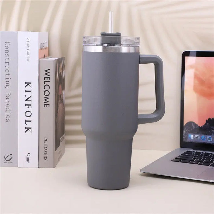 Grå rostfritt stål kaffemugg med handtag och sugrör bredvid bok och laptop på skrivbord