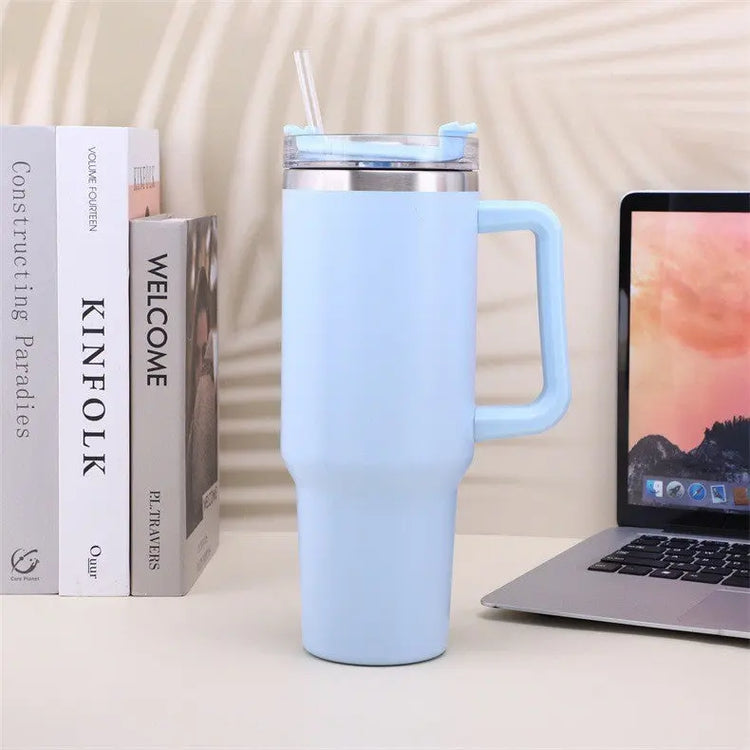 Ljusblå rostfritt stål kaffemugg med handtag och genomskinligt lock på skrivbord bredvid laptop och böcker